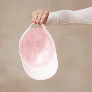 Op de afbeelding: Een witte baseballcap met een roze binnenkant wordt vastgehouden. De cap heeft een gebogen klep en een stoffen band. De binnenkant is zachtroze en de buitenkant is wit.