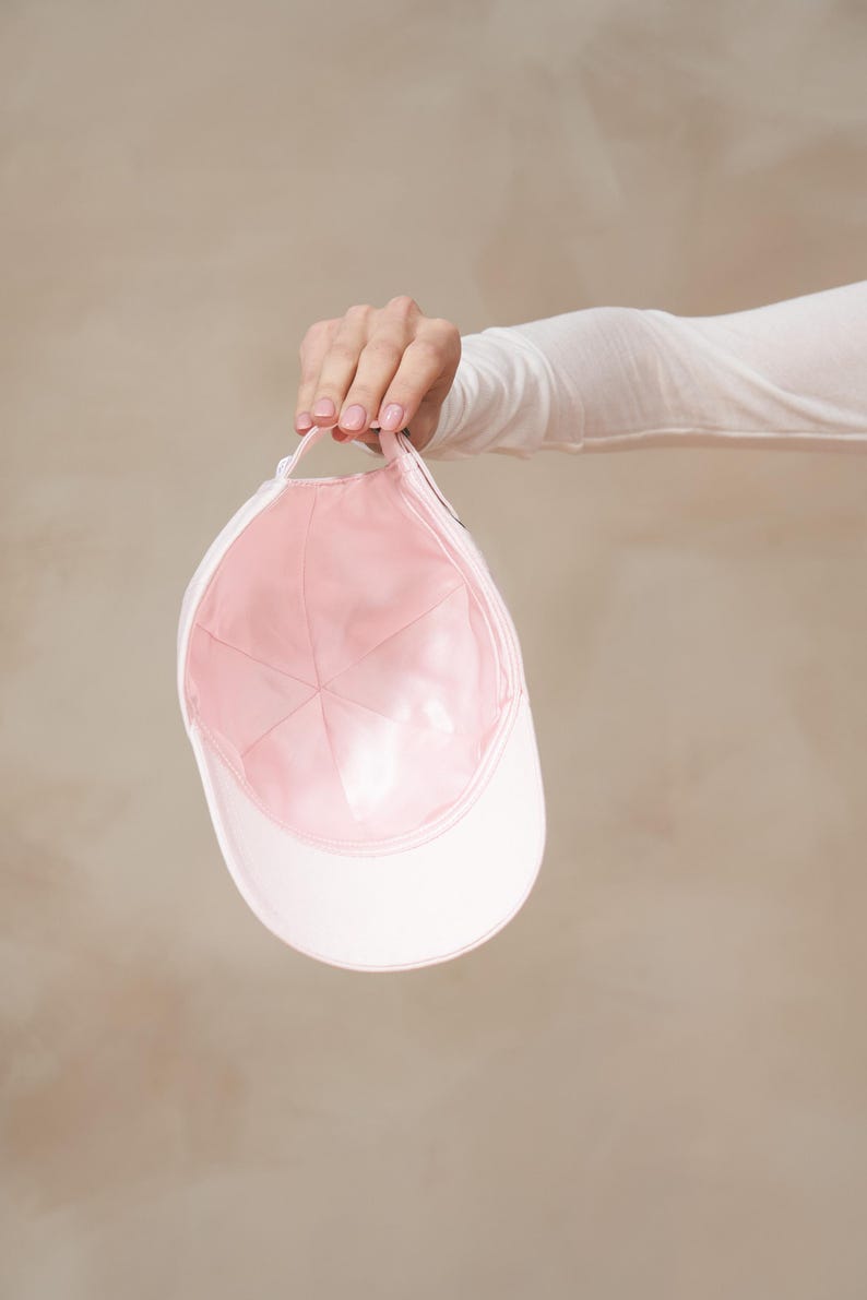 Op de afbeelding: Een witte baseballcap met een roze binnenkant wordt vastgehouden. De cap heeft een gebogen klep en een stoffen band. De binnenkant is zachtroze en de buitenkant is wit.