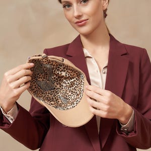 Op de afbeelding: Een beige baseballpet met een luipaardprint voering. De pet wordt omhoog gehouden om het binnenpatroon te laten zien. De persoon op de foto draagt een bordeaux blazer en een cr&egrave;mekleurige blouse.