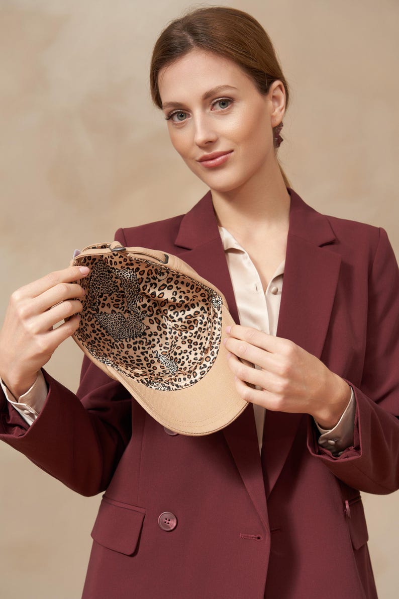 Op de afbeelding: Een beige baseballpet met een luipaardprint voering. De pet wordt omhoog gehouden om het binnenpatroon te laten zien. De persoon op de foto draagt een bordeaux blazer en een cr&egrave;mekleurige blouse.