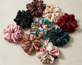 May include: An assortment of satin scrunchies in various colors and patterns. Colors include black, brown, burgundy, teal, pink floral, cream floral, and green. The scrunchies are arranged in a circular pattern on a neutral background.