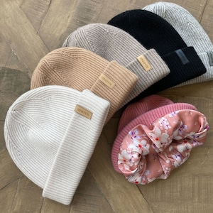 May include: A collection of six knitted beanies in various colours, including grey, black, beige, and pink. Each beanie has a small leather tag with the word "Alanna" on it. The beanies are arranged in a semi-circle on a wooden surface.