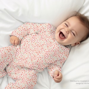 May include: A baby wearing a pink and white floral print onesie, lying on a white sheet, smiling and looking up.