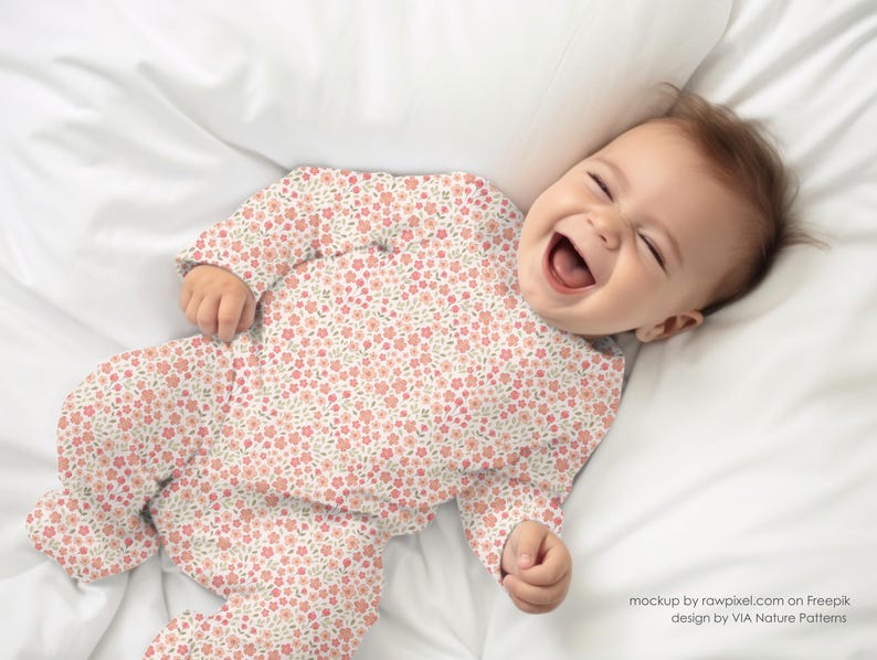 May include: A baby wearing a pink and white floral print onesie, lying on a white sheet, smiling and looking up.