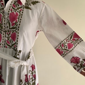 May include: White cotton dress with a floral print in pink and green. The dress features long sleeves, a collared neckline, and a matching belt. The floral pattern is repeated on the sleeves and bodice.