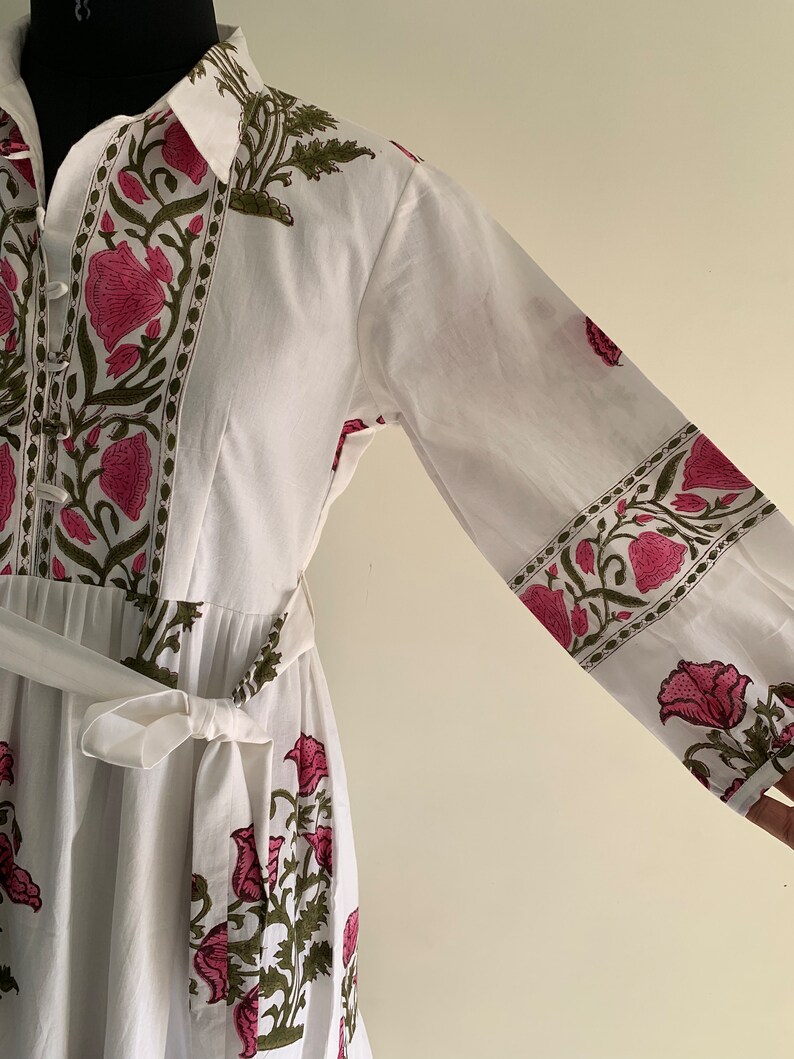 May include: White cotton dress with a floral print in pink and green. The dress features long sleeves, a collared neckline, and a matching belt. The floral pattern is repeated on the sleeves and bodice.