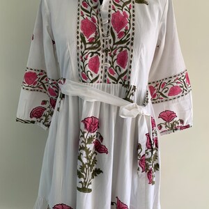May include: White dress with a floral print in pink and green. The dress features a collared neckline, button-down front, and a tie belt at the waist. The sleeves are three-quarter length, and the skirt has a tiered, ruffled hem.