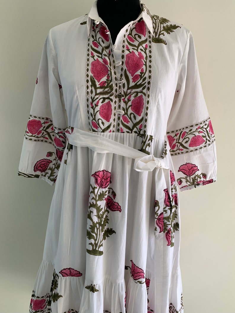 May include: White dress with a floral print in pink and green. The dress features a collared neckline, button-down front, and a tie belt at the waist. The sleeves are three-quarter length, and the skirt has a tiered, ruffled hem.