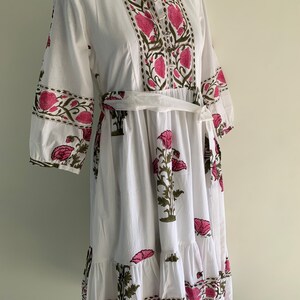 May include: White cotton dress with a floral print in pink and green. The dress features a collared neckline, three-quarter sleeves, and a matching belt. The floral pattern is repeated on the sleeves and the bottom hem.
