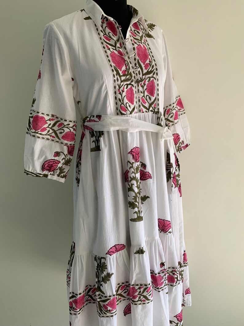May include: White cotton dress with a floral print in pink and green. The dress features a collared neckline, three-quarter sleeves, and a matching belt. The floral pattern is repeated on the sleeves and the bottom hem.
