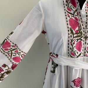 May include: White dress with a floral pattern in pink and green. The dress features long sleeves, a collared neckline, and a matching belt. The floral design is printed along the sleeves, neckline, and the front of the dress.
