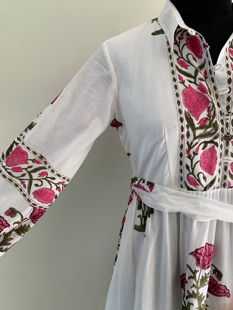 May include: White dress with a floral pattern in pink and green. The dress features long sleeves, a collared neckline, and a matching belt. The floral design is printed along the sleeves, neckline, and the front of the dress.