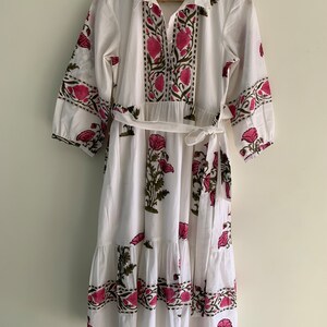 May include: White midi dress with a floral print in pink and green. The dress features a collared neckline, button-down front, and a matching belt. The sleeves are three-quarter length.