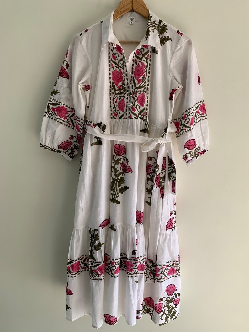 May include: White midi dress with a floral print in pink and green. The dress features a collared neckline, button-down front, and a matching belt. The sleeves are three-quarter length.