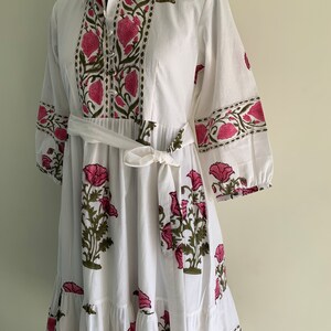 May include: White cotton dress with a floral print in pink and green. The dress features a collared neckline, button-down front, and a tie belt at the waist. The sleeves are three-quarter length, and the skirt has a tiered design.