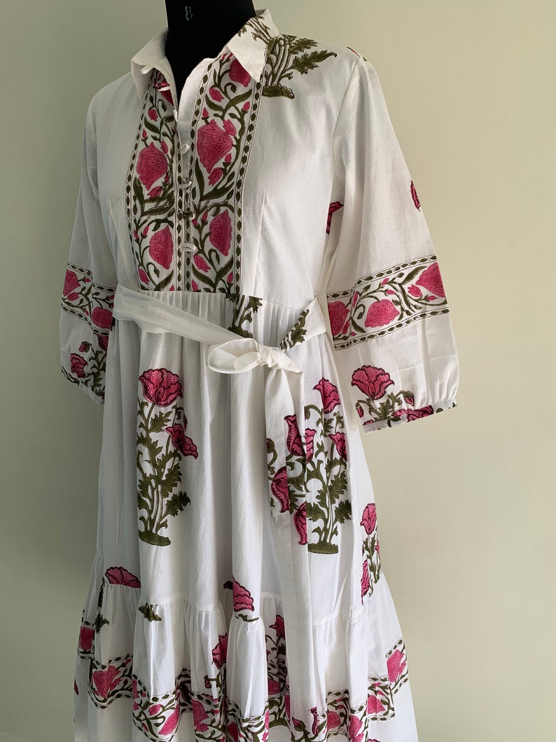 May include: White cotton dress with a floral print in pink and green. The dress features a collared neckline, button-down front, and a tie belt at the waist. The sleeves are three-quarter length, and the skirt has a tiered design.