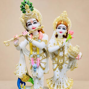 May include: A white statue of two figures standing on a rectangular base. The left figure holds a gold flute and wears a peacock-feather crown. The right figure holds a pink flower and a gold pot. Both wear white garments with gold accents.