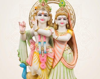 May include: A white marble statue of Radha and Krishna standing together. Krishna plays a flute, wearing a yellow dhoti and crown with a peacock feather. Radha wears a peach and green sari. A small blue peacock stands at their feet on a base.