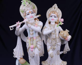 May include: White marble statues of a male figure playing a flute and a female figure holding a flower and a pot. Both figures are adorned with gold accents and elaborate headdresses. The statues stand on a decorative base.