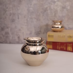 May include: A shiny, gold-coloured metal vase with a rounded body and a flared rim. The vase is sitting on a white surface.