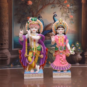 Puede incluir: Un par de estatuas coloridas pintadas a mano de deidades hind&uacute;es, Krishna y Radha, de pie sobre bases separadas. Krishna sostiene una flauta y Radha sostiene una flor de loto. Las estatuas est&aacute;n decoradas con intrincados detalles y se colocan sobre un fondo de dise&ntilde;o de plumas de pavo real.