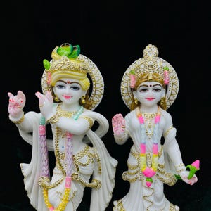 May include: White marble statue of Radha Krishna, a Hindu deity couple, with intricate details and colorful accents. The statues are standing on separate bases with ornate designs.
