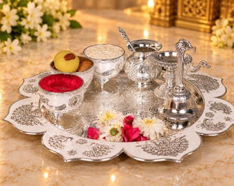 May include: A silver-toned metal puja thali set featuring a scalloped tray with intricate peacock engravings. The set includes small bowls containing red powder and rice, a small pot with a spoon, and a bell with a peacock-shaped handle.