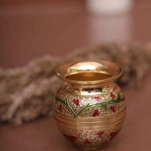 Puede incluir: Una peque&ntilde;a olla de metal dorada con intrincados dise&ntilde;os florales rojos y verdes. La olla est&aacute; decorada con un borde elevado y tiene un acabado liso y pulido.