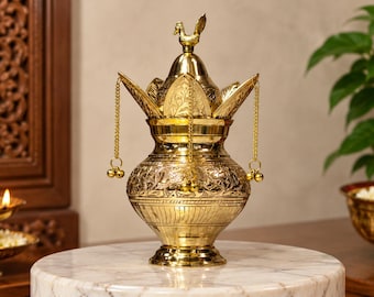 May include: A polished brass kalash vessel featuring intricate floral engravings on the body and a lid topped with a small bird figurine. Three delicate gold-toned chains with small spherical bells hang from the lid's pointed petal-like edges.