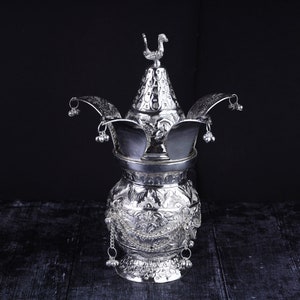 May include: A silver ceremonial pot with intricate floral designs and a bird finial on the lid. The pot has a round body and a wide base, and is decorated with bells and chains.