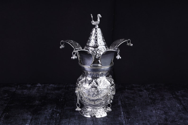 May include: A silver ceremonial pot with intricate floral designs and a bird finial on the lid. The pot has a round body and a wide base, and is decorated with bells and chains.