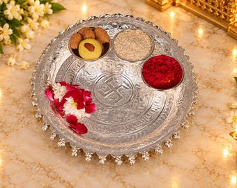 May include: A round silver-toned metal plate with an embossed swastika symbol in the center and scalloped edges. The plate holds two small bowls containing grains and red powder, along with a cluster of pink and white flower petals.
