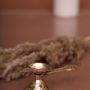 Puede incluir: Una peque&ntilde;a l&aacute;mpara de aceite dorada con una base decorativa y un asa. La base est&aacute; decorada con motivos florales verdes y rojos.
