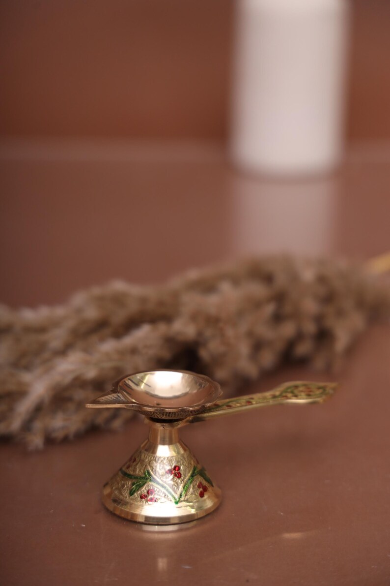 Puede incluir: Una peque&ntilde;a l&aacute;mpara de aceite dorada con una base decorativa y un asa. La base est&aacute; decorada con motivos florales verdes y rojos.