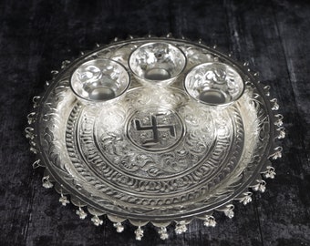May include: A silver serving tray with three small bowls and an engraved swastika symbol in the center. The tray has a decorative border and hanging bells.