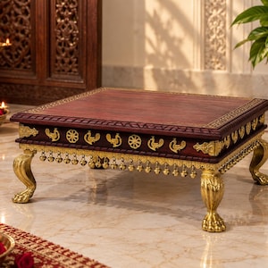 May include: A rectangular wooden table with a dark brown top and gold-coloured metal accents. The sides feature repeating bird and wheel motifs, while the base is supported by four ornate, claw-shaped legs with hanging metal bells along the edge.