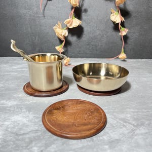 Op de afbeelding: Een set messing serveerartikelen, waaronder een kleine beker met een lepel, een kom en een houten onderzetter met een bloemenontwerp. De items zijn gerangschikt op een grijze ondergrond, met bloemendecoraties op de achtergrond.