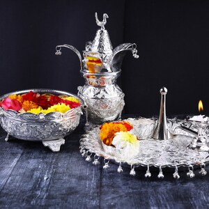 May include: A silver ceremonial set with a decorative pitcher, a bowl filled with colorful flowers, and a tray with a lit candle and a silver diya.