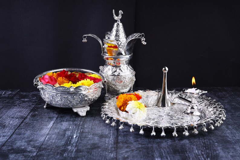 May include: A silver ceremonial set with a decorative pitcher, a bowl filled with colorful flowers, and a tray with a lit candle and a silver diya.