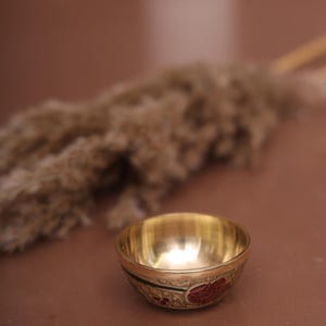 Puede incluir: Un peque&ntilde;o cuenco redondo de color dorado con intrincados dise&ntilde;os rojos y verdes. El cuenco est&aacute; sobre una superficie marr&oacute;n.