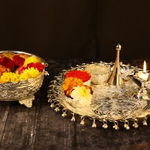 May include: A silver tray with a decorative design, holding a small bowl, a silver pitcher, a lit candle, and a bowl filled with yellow, orange, and red flowers.