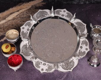 May include: A silver-colored serving tray with a detailed peacock design, surrounded by smaller decorative bowls. The tray is round with a scalloped edge, and the bowls contain various food items. The arrangement is set on a purple surface.