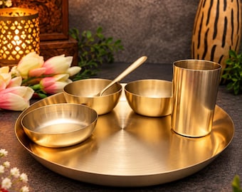 May include: A polished brass dinnerware set consisting of a large round serving tray, two small bowls, a tall cylindrical tumbler, and a matching spoon. The items feature a smooth, metallic gold finish with a clean, minimalist circular design.