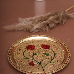 Puede incluir: Una bandeja redonda de metal dorada con un dise&ntilde;o decorativo de flores rojas y hojas verdes. La bandeja tiene un borde elevado y una superficie texturizada.
