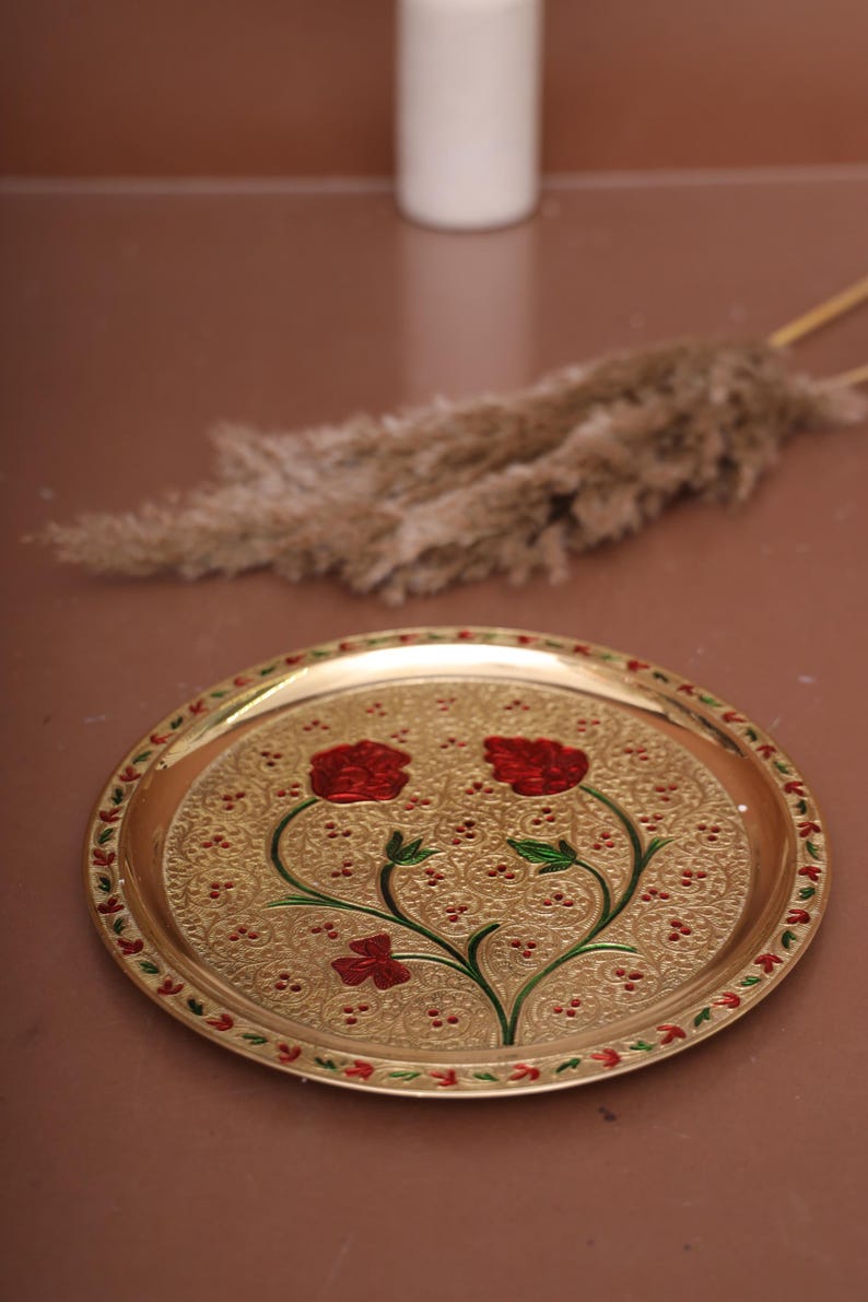 Puede incluir: Una bandeja redonda de metal dorada con un dise&ntilde;o decorativo de flores rojas y hojas verdes. La bandeja tiene un borde elevado y una superficie texturizada.