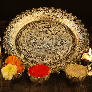 May include: A silver tray with an ornate design featuring two peacocks. The tray is surrounded by small bowls filled with colorful powders and flowers. A small oil lamp with a lit wick is also visible.