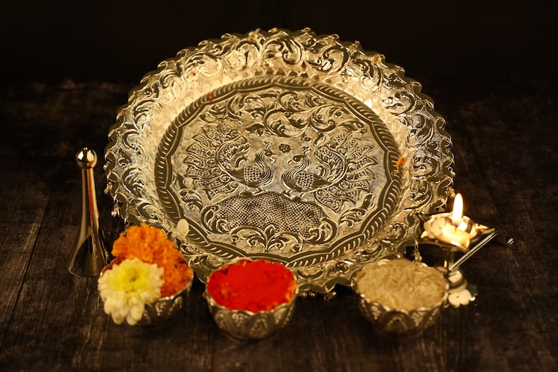 May include: A silver tray with an ornate design featuring two peacocks. The tray is surrounded by small bowls filled with colorful powders and flowers. A small oil lamp with a lit wick is also visible.