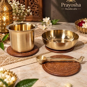 May include: A set of brass kitchenware including a cylindrical cup, a shallow bowl, and a long-handled spoon, each resting on a circular dark brown wooden coaster with carved floral patterns. The brand name Prayosha Handicrafts is visible.