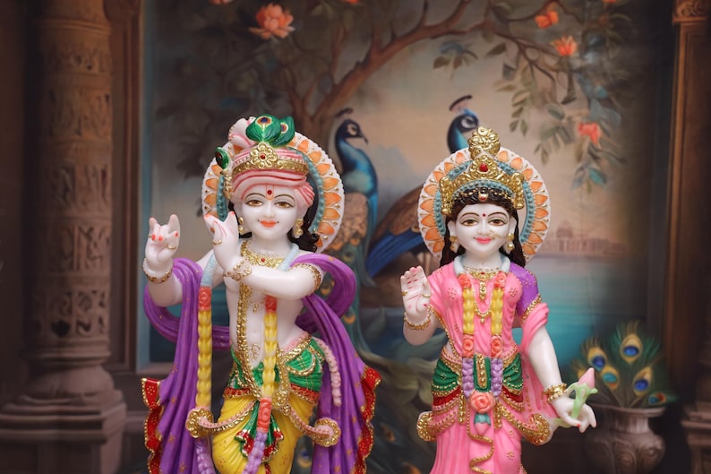 Puede incluir: Una estatua de m&aacute;rmol blanco de Krishna y Radha, una pareja de deidades hind&uacute;es, de pie juntos. Krishna sostiene una flauta y Radha sostiene una flor de loto. Las estatuas est&aacute;n decoradas con pintura colorida y joyas.