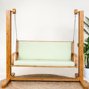 Può includere: Un dondolo in legno con seduta e schienale imbottiti color verde chiaro, sospeso tramite cavi metallici a una robusta struttura in legno. Il telaio presenta due montanti verticali collegati da una base che sostiene la panca sospesa.
